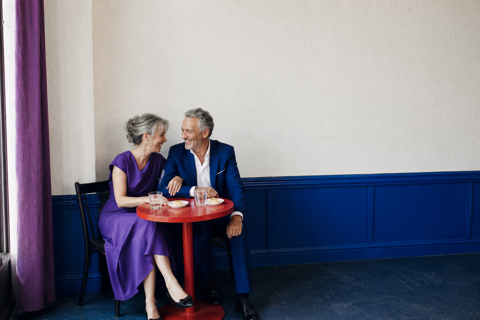 Pareja adulta conversa y sonríe alrededor de una mesa roja en un espacio azul y violeta
