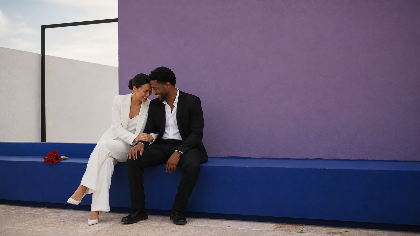 Pareja sentada se acerca y sonríe frente a un muro violeta y una banca azul