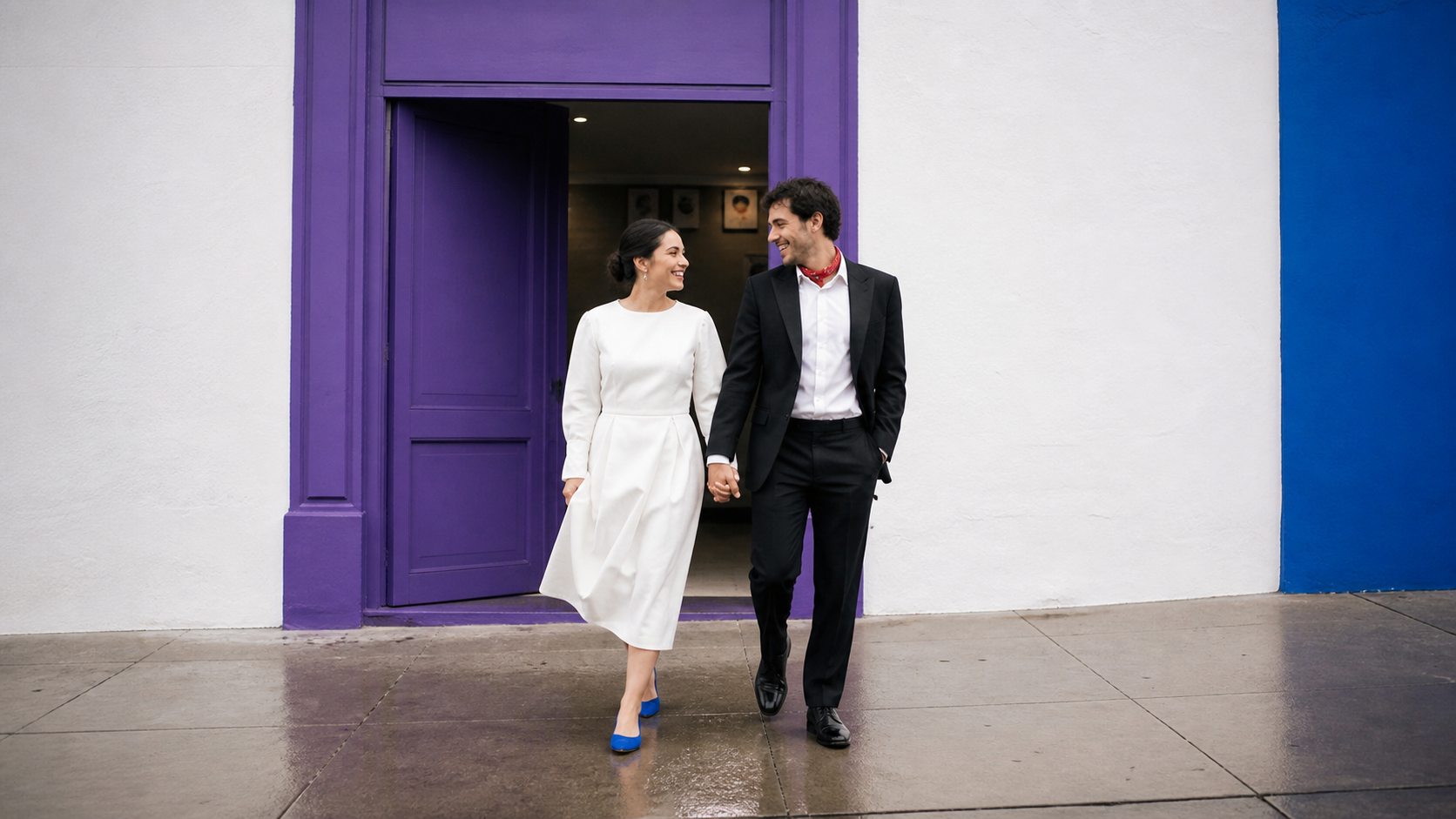 Pareja vestida de blanco y negro sale de una puerta violeta y camina de la mano frente a una fachada marfil y azul