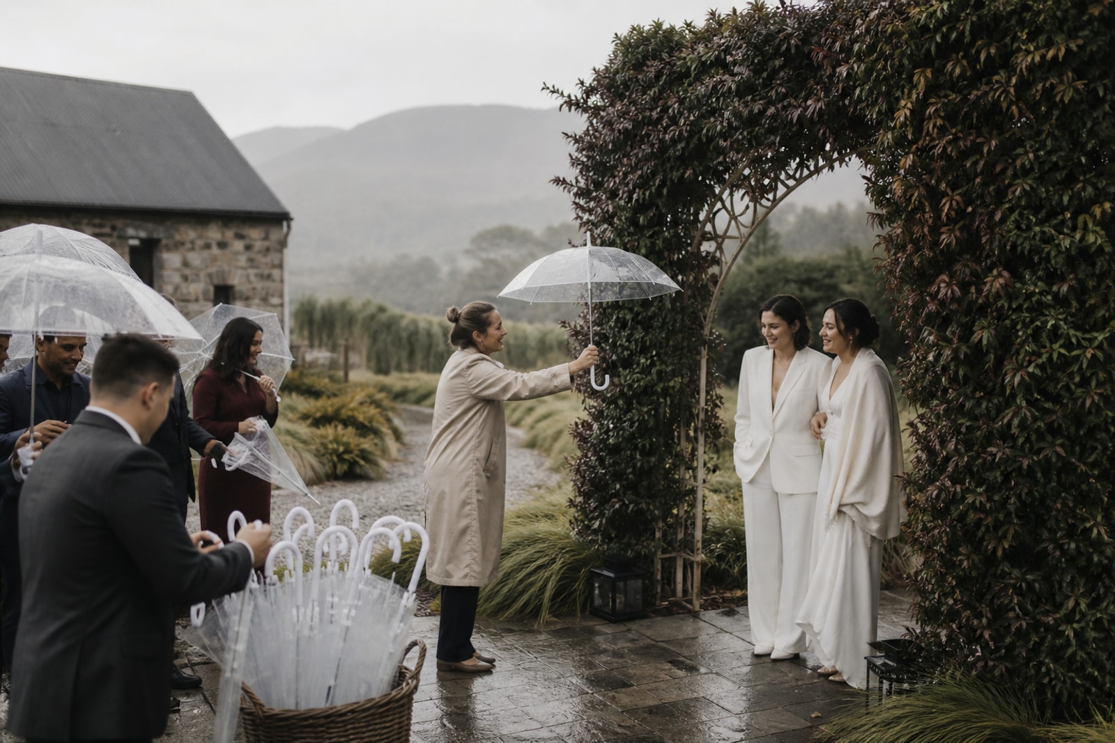 Una wedding planner reparte paraguas y la pareja avanza tranquila por un paso cubierto.