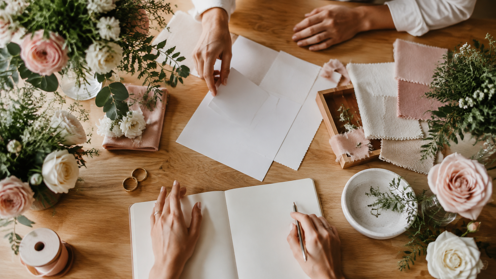 Dos personas colaboran en una mesa de planeación de boda con flores, anillos, telas y papelería en blanco.